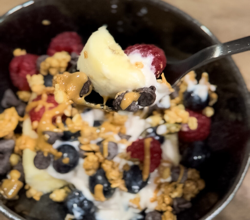 Spoon lifting a bite of banana, yogurt, raspberry, and chocolate chips from a bowl of breakfast sundae.