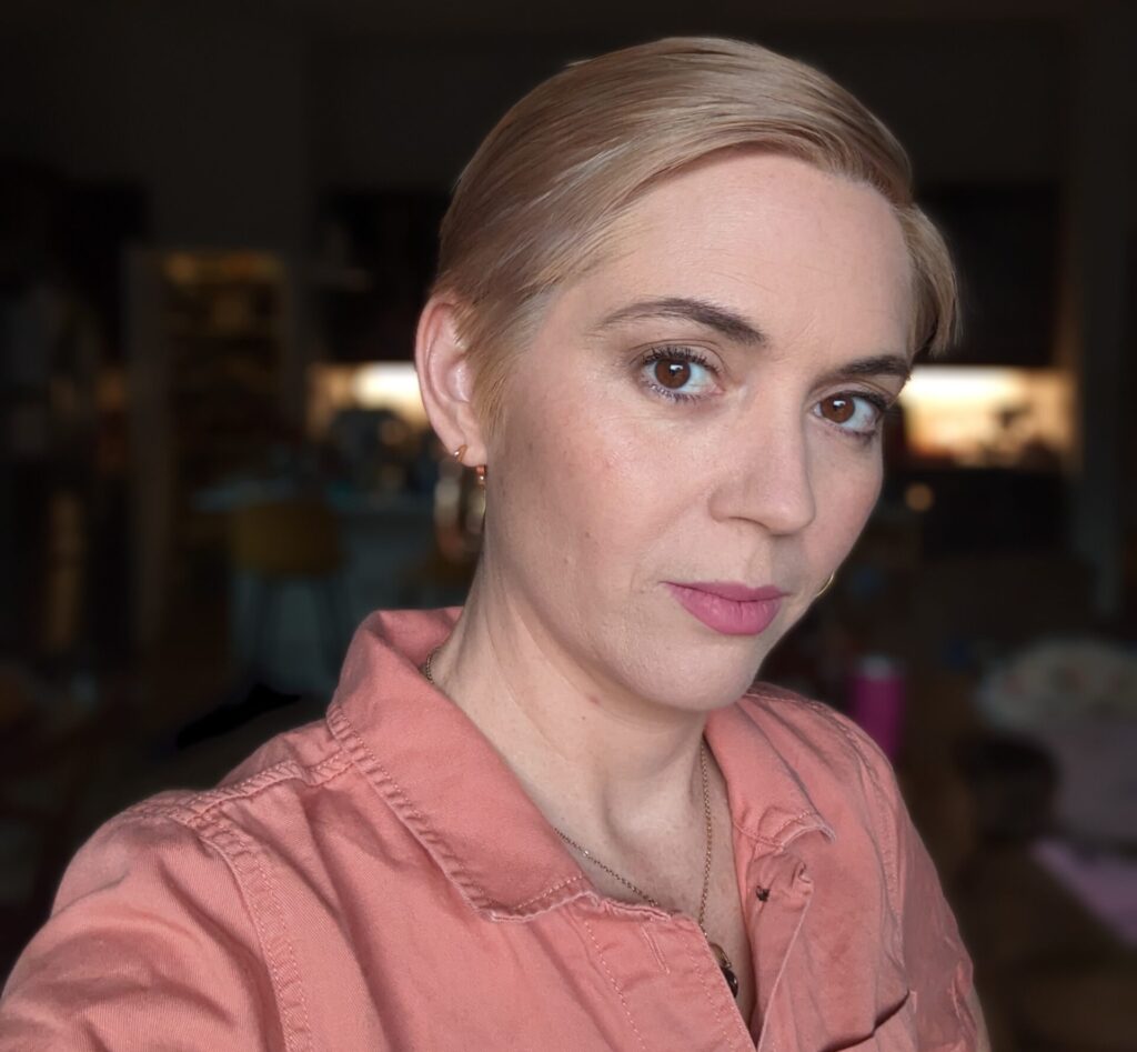 Lauren wearing a coral button-down shirt, smiling softly in front of a dimly lit kitchen. She has short platinum-blonde hair and gold hoop earrings.