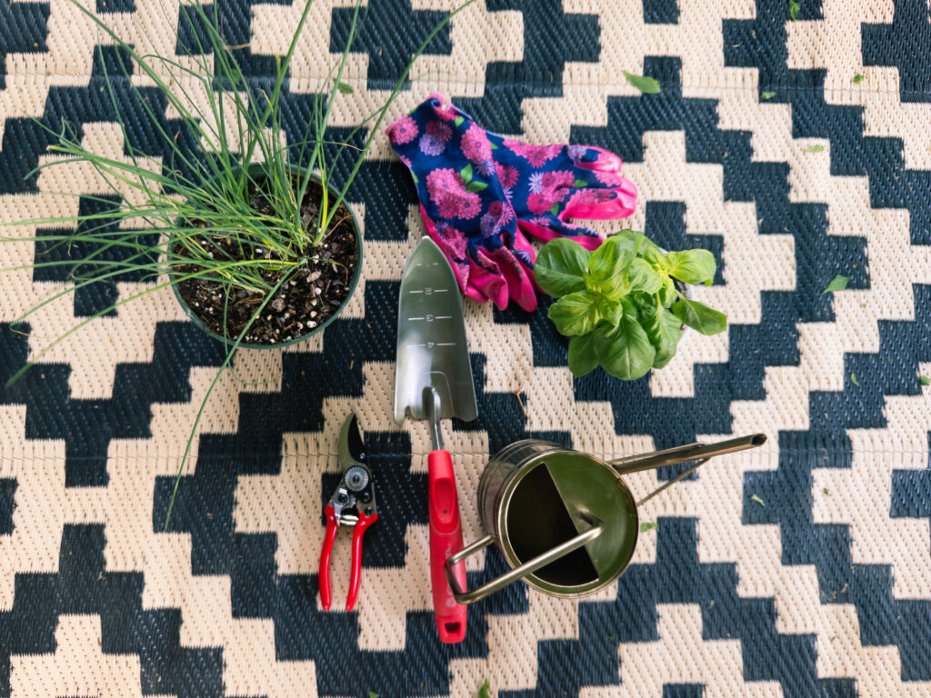 Flat lay of herb gardening tools, chives, basil, and gloves on a patterned patio rug